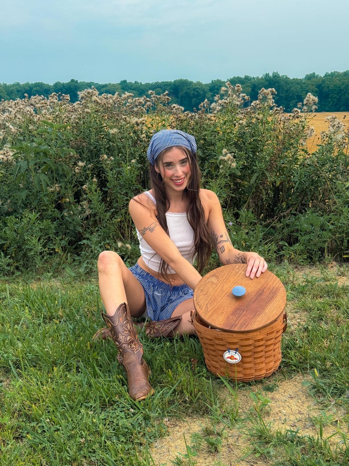 Livinia smiling in a blue gingham headscarf, sitting in the grass with a picnic basket